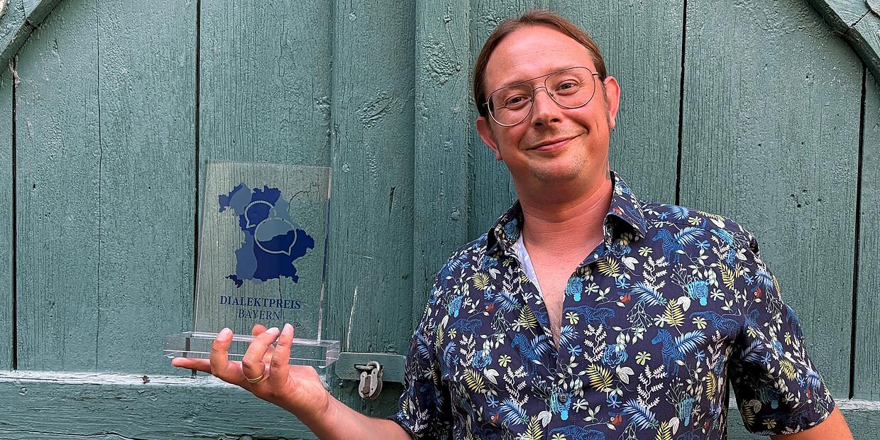 Wastel Kauz hält lächelnd die gläserne Trophäe des Dialektpreises Bayern vor einer grünen Holztür